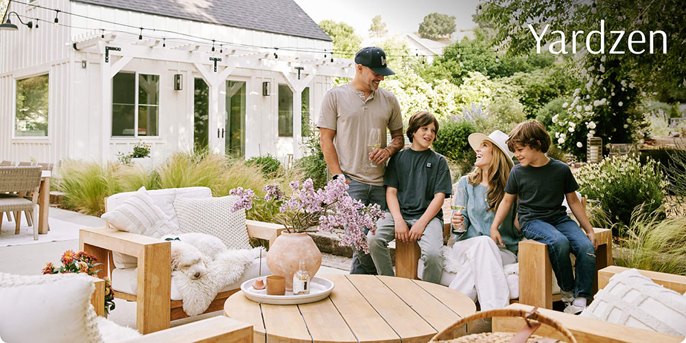 A man and three children sit and chat on a stylish patio surrounded by greenery and cozy wooden furniture, with a white house and string lights in the background. The word Yardzen appears in the top right corner.