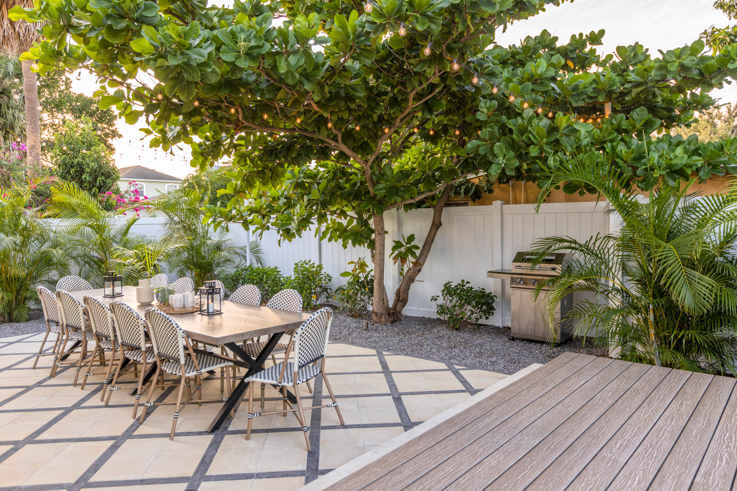 A backyard patio with a long dining table and chairs under a large leafy tree, string lights, potted plants, a barbecue grill, and a white fence surrounding the area.