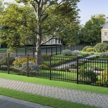A well-maintained garden with neatly trimmed shrubs and a large tree is enclosed by a black metal fence. A stone walkway and a pathway run through green grass, and houses are visible in the background among trees.