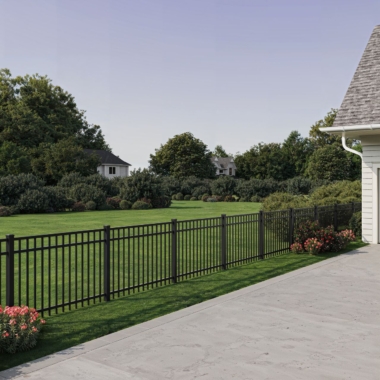 A black metal fence separates a neatly manicured lawn from a concrete patio next to a house, with bushes and trees in the background under a clear sky.