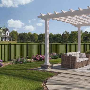 A backyard patio with a white pergola, wicker outdoor sofa, and landscaped garden, overlooking a grassy yard with a black metal fence and a house in the distance, surrounded by trees under a blue sky.