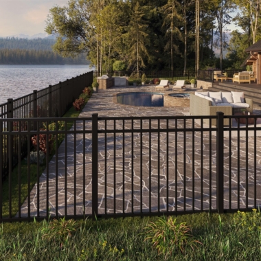 A black metal fence surrounds a patio area with stone paving, outdoor furniture, and a pool, set beside a lake and bordered by trees in a peaceful, natural landscape.