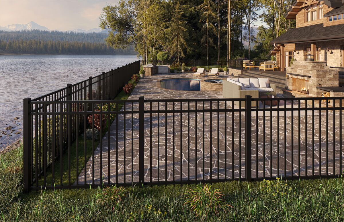 A black metal fence encloses a stone patio with outdoor seating and a pool, beside a large house by a lake, surrounded by trees and mountains in the background.