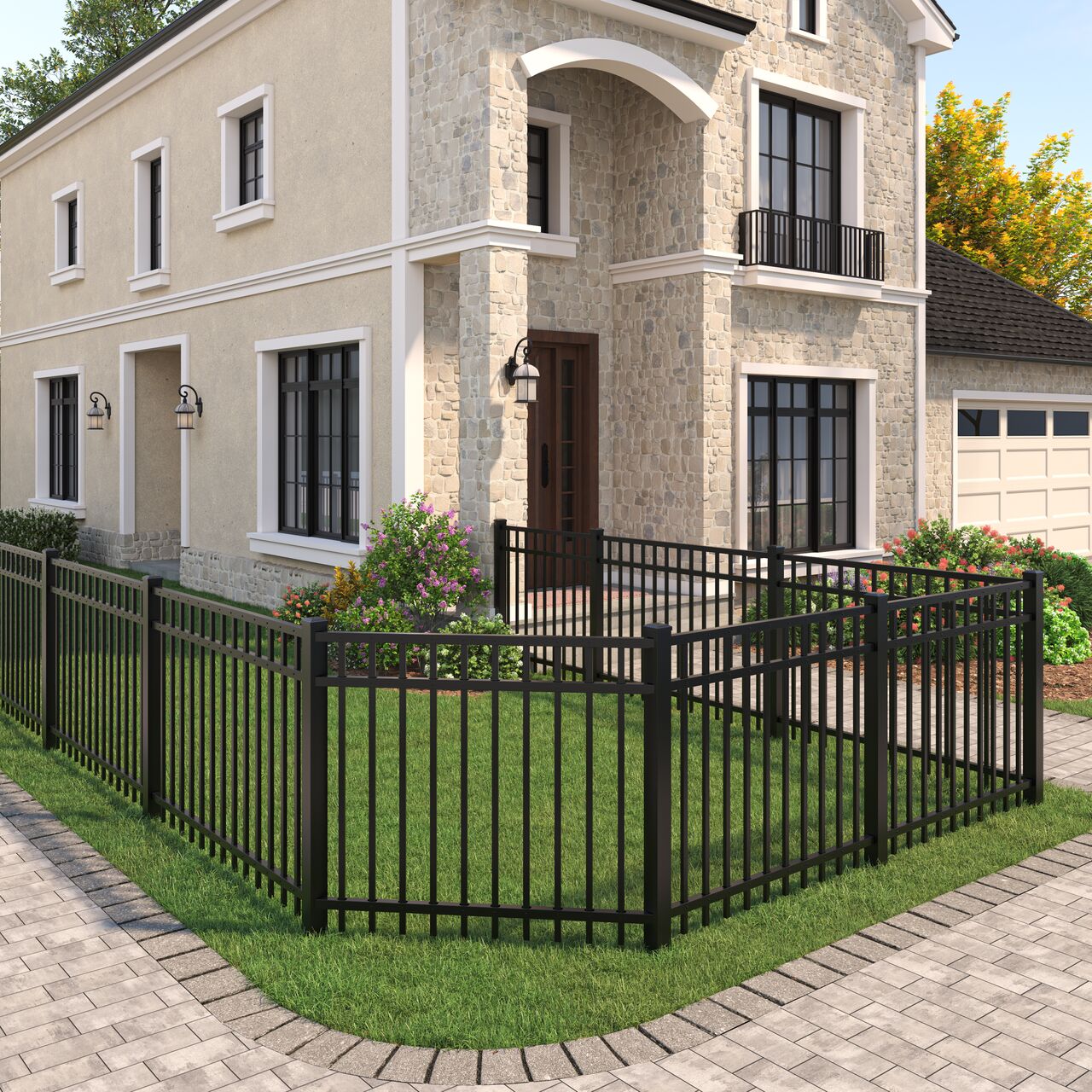 A beige stone house with dark trim and a small balcony, surrounded by a black metal fence enclosing a green lawn with shrubs and flowers. A brick driveway leads to a garage on the right.