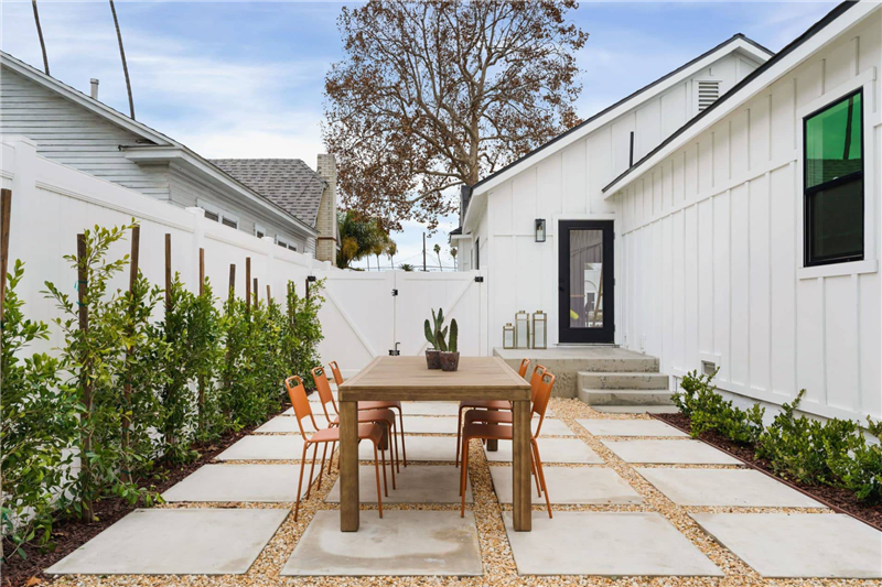 A modern backyard patio with a wooden dining table and eight orange chairs sits on large square pavers surrounded by small gravel. Green shrubs line the white fence, and a white house stands in the background.