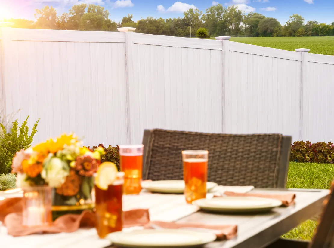 A sunlit outdoor table is set with plates and drinks. A bouquet of flowers decorates the table, and a white fence with green fields and trees is in the background.