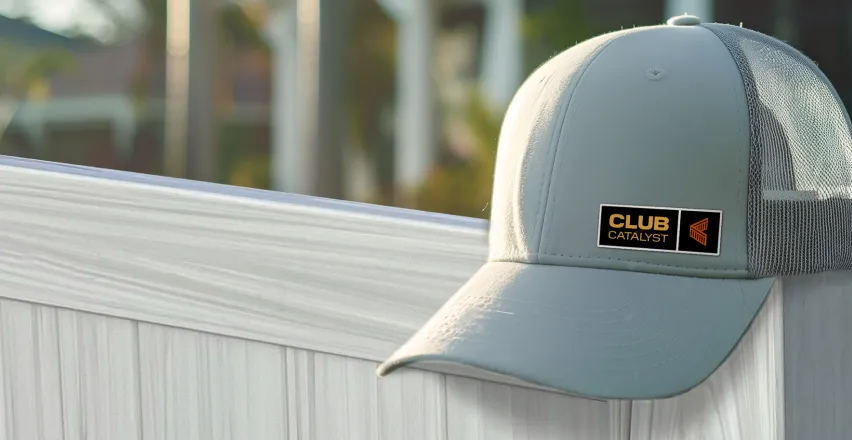 A light gray baseball cap with a black and gold Club Catalyst patch rests on a white wooden fence. The background is blurred with hints of greenery and sunlight.