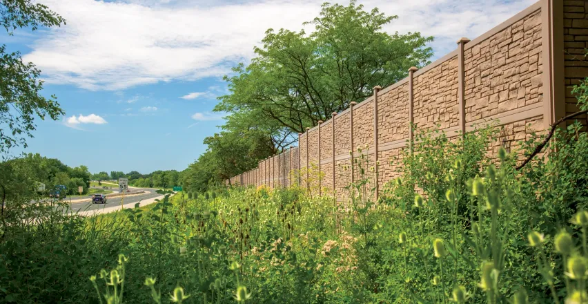 A tall, textured concrete wall lines a highway surrounded by green vegetation and trees. The sky is blue with scattered clouds. A car is visible driving on the road in the distance.